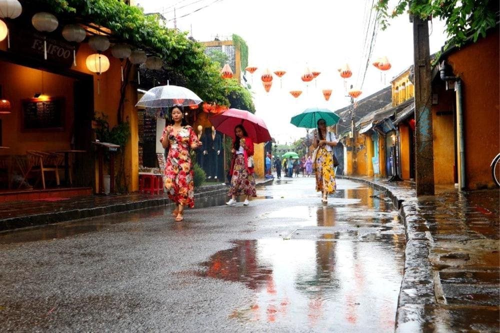 Locals and travelers take shelter under shop awnings as warm rain falls on a quiet Hoi An street